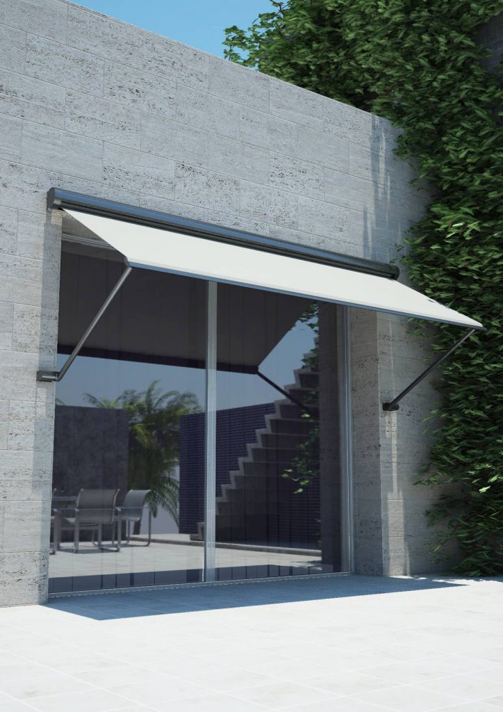 A modern, beige retractable awning extends over a glass sliding door on a stone building with greenery climbing one side.