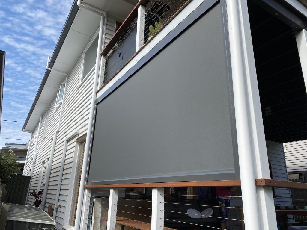 A gray outdoor blind covers the balcony area of a two-story house with white siding, seen from the exterior against a blue sky.