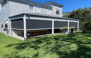 A two-story house with an extended patio featuring drop-down shades, wooden dining tables, and chairs underneath. The backyard is grassy with potted plants and a black dog in the shade.