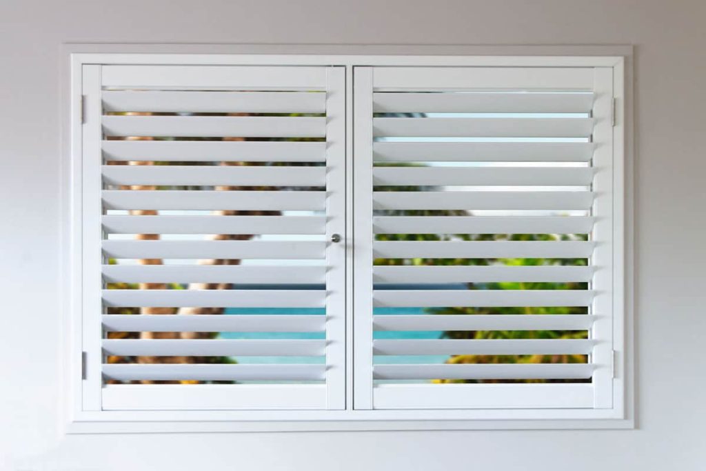 A closed white window with horizontal aluminium shutters reveals a blurry view of a tropical scene outside.