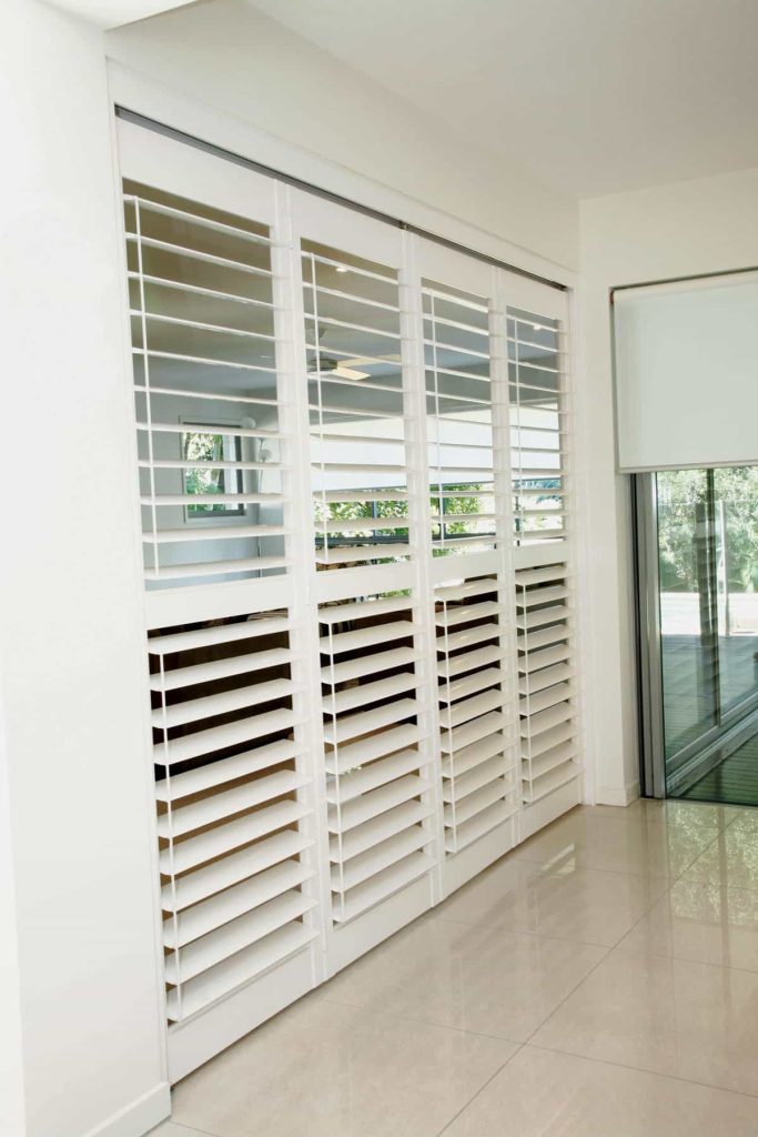 Room with large, white PVC shutters covering wide windows, allowing partial view outside. Adjacent glass door leads to an outdoor area. Floor is tiled in a light color.