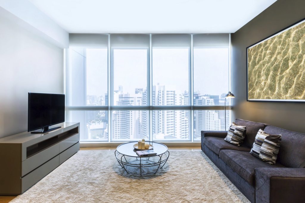Modern living room with large windows featuring internal blinds offering a city view, a dark sofa with patterned cushions, a wireframe coffee table, a TV on a low cabinet, and a framed wall art piece.