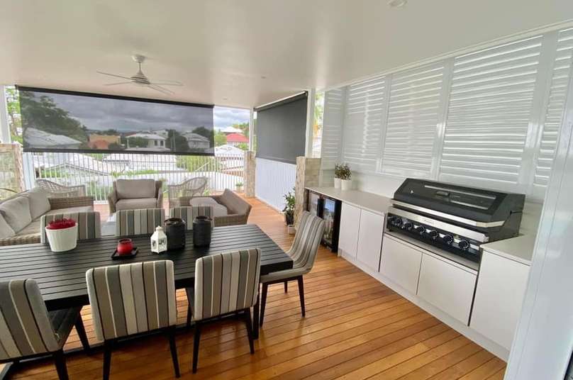 A covered outdoor patio with a dining table and chairs, a seating area with sofas, a barbecue grill, and a ceiling fan. The space features wood flooring, white privacy shutters, and Outdoor Blinds Brisbane for added comfort.