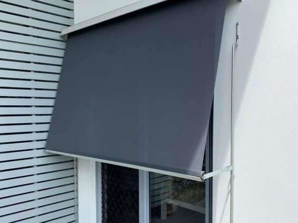 A window with a dark gray retractable awning partially extended, mounted on a white wall next to a set of white horizontal slat panels, provides an elegant touch often seen in homes that feature Outdoor Blinds Brisbane.