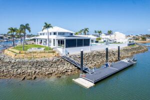 A waterfront house with a white roof is situated on a rocky shoreline. It features a modern design, a small backyard with a pool, and a private dock extending into the calm water. Palm trees and neighboring homes are visible in the background.