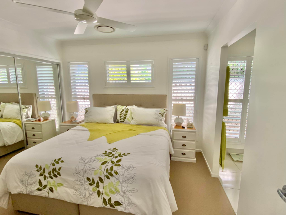 A cozy bedroom with a double bed covered in a white and green floral duvet. The room features mirrored closets, bedside tables with lamps, and a ceiling fan. Indoor PVC shutters adorn the windows, allowing natural light to filter through and create a warm ambiance.