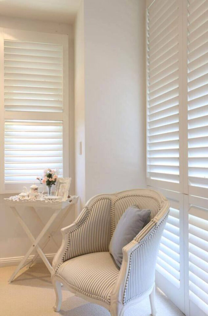 A cozy corner with a striped armchair and a side table adorned with flowers and photo frames. The room features large windows with Indoor PVC Shutters, allowing soft natural light to filter in. The decor remains elegant and tranquil.