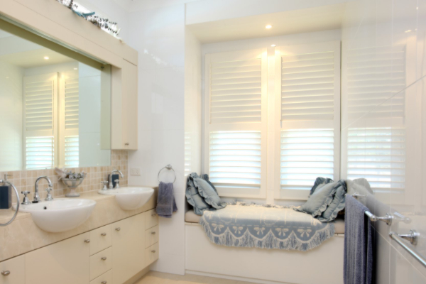 A modern bathroom with twin sinks and a large mirror on the left. A cozy window seat with blue cushions and a patterned throw invites relaxation. Indoor PVC shutters cover the window, complementing towels hanging from racks. The decor features soft, neutral tones.