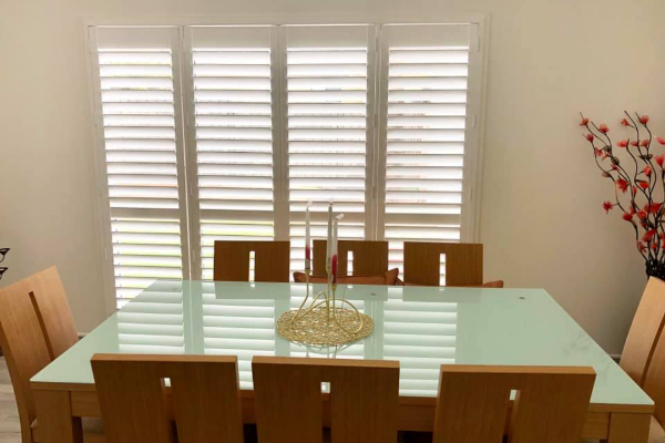 A dining room with a glass-topped table surrounded by six wooden chairs. Behind the table, there are white indoor PVC shutters. A decorative centerpiece adorns the table, while red ornamental branches stand elegantly in a vase on the right side.