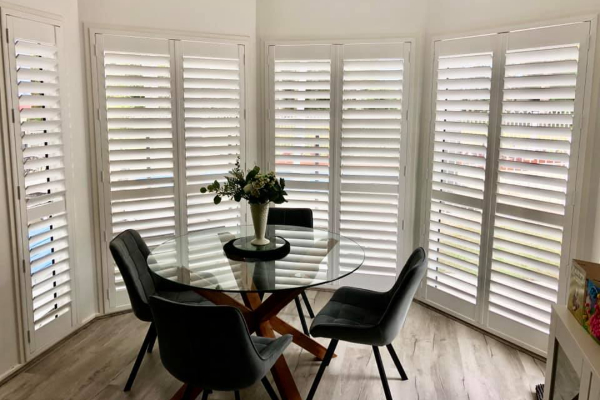 A dining area features a round glass table and four dark chairs, surrounded by white indoor PVC shutters on large windows. These shutters allow diffused light to fill the space. A vase with flowers is centered on the table, adding a touch of elegance.