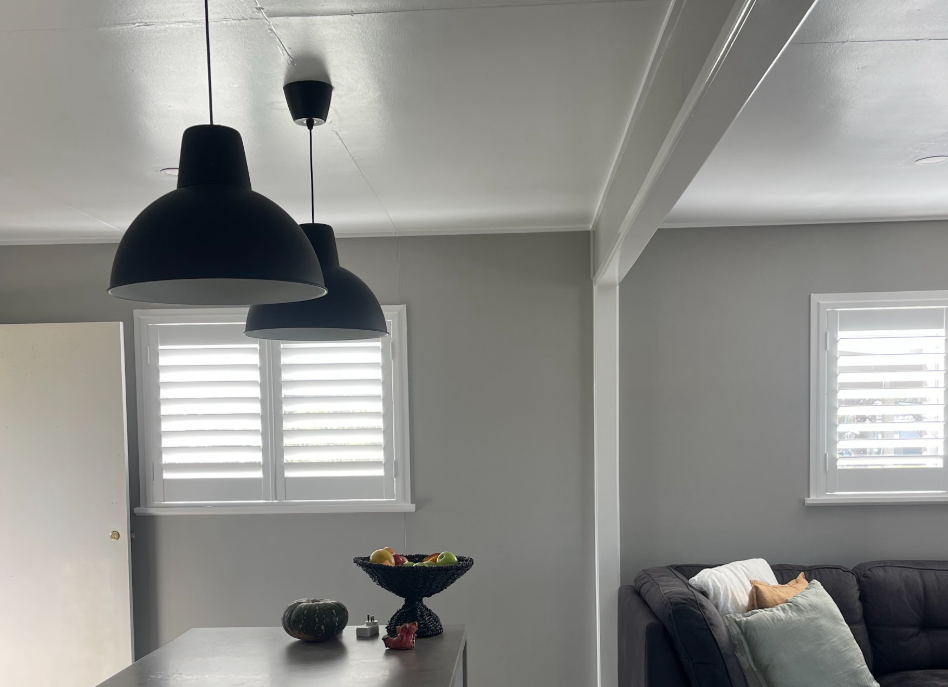 A modern living room with gray walls and a sofa adorned with cushions. Suspended black pendant lights hang from the ceiling. A fruit bowl and a small pumpkin sit on a countertop. White indoor PVC shutters allow natural light in, enhancing the room's elegance.