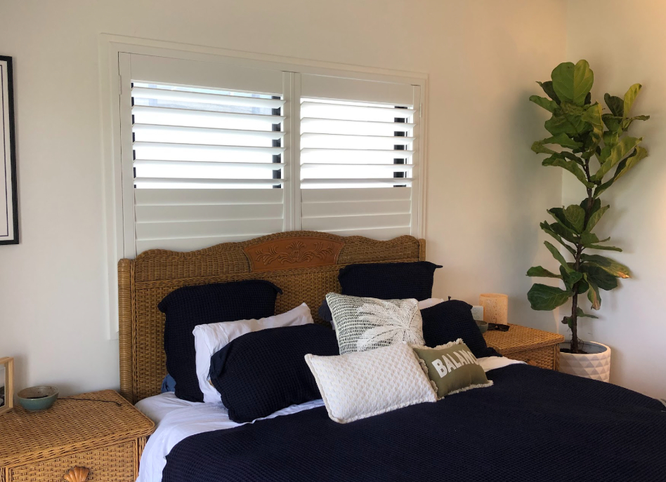 A cozy bedroom features a wicker headboard and matching side tables. The bed is dressed in navy and white linens with pillows, including one that says "BALM." A tall, leafy plant stands by the bed beneath sleek indoor PVC shutters.