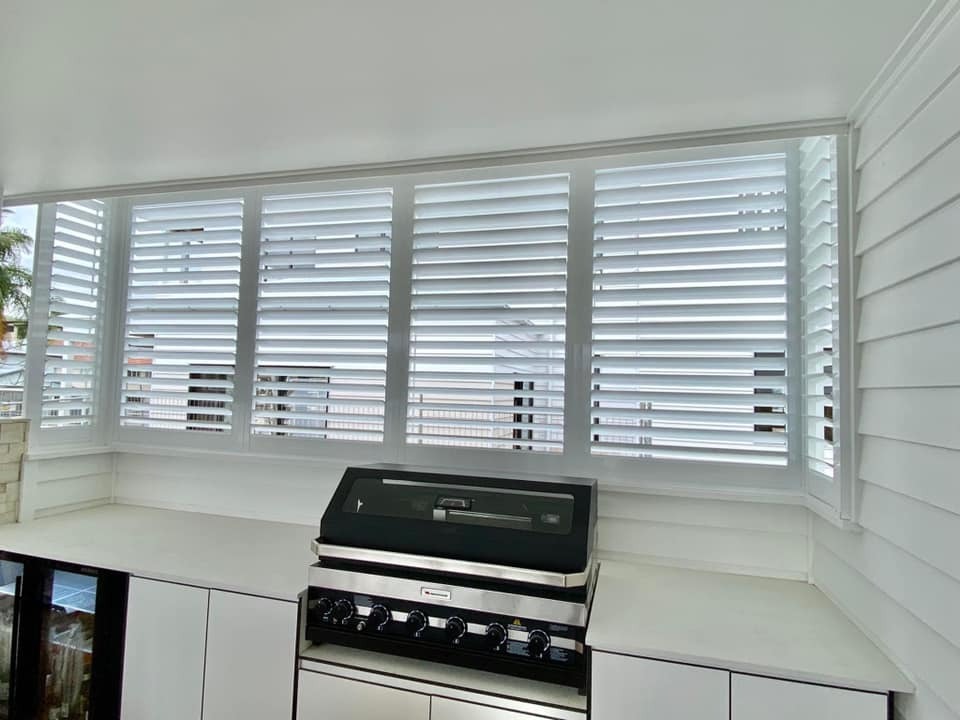 A modern indoor kitchen features a sleek countertop with a stainless steel grill. Large outdoor aluminium shutters on the window allow natural light to filter in, enhancing the clean, minimalist design.