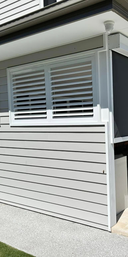 A modern exterior wall with horizontal paneling highlights a window adorned with sleek white outdoor aluminum shutters. The area below the window is painted gray, complemented by a concrete walkway and lush green grass in the foreground.