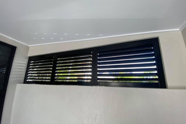 A horizontal row of black, outdoor aluminium shutters near the ceiling allows natural light to filter into the room. The walls are painted light beige and the ceiling is white, creating a modern and minimalist interior space.