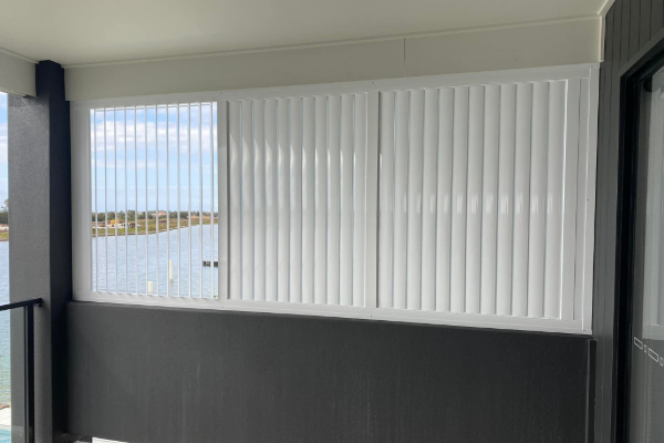 A balcony view with vertical white blinds, paired with sleek outdoor aluminium shutters, partially closed. They overlook a calm body of water under a cloudy sky, casting gentle shadows on the floor beneath. The scene conveys a peaceful and serene atmosphere.