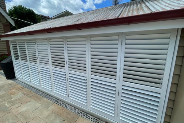 Outdoor aluminium louvered shutters adorn the side of a building with a corrugated metal roof. The ground is paved with light-colored tiles, and a trash bin sits by the building under a partly cloudy sky.