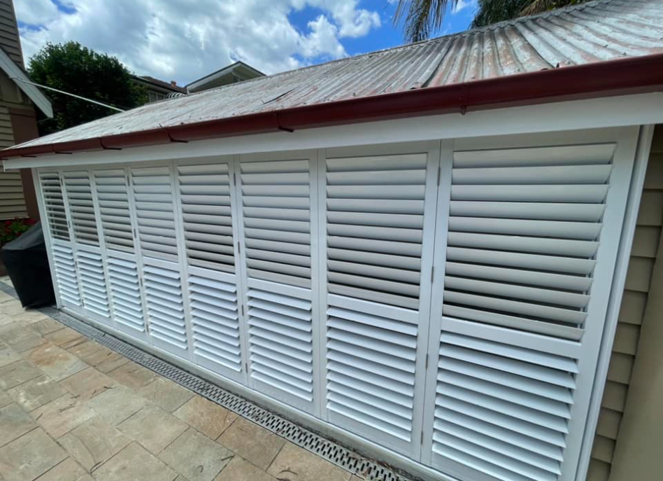 A row of white outdoor aluminium shutters covers a long window on a building with a corrugated metal roof. The background showcases a cloud-filled blue sky with glimpses of other structures, while the ground is paved with stone tiles.