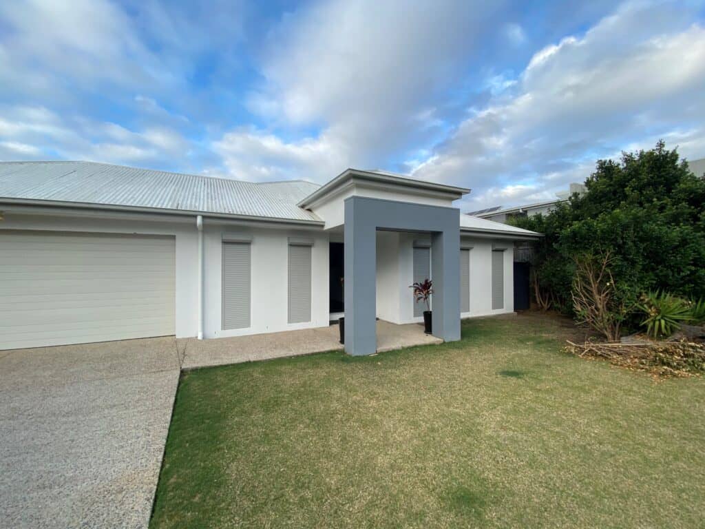 Modern single-story house with a grey and white exterior, a covered entryway, roll-down window shutters, and a small lawn with minimal landscaping.