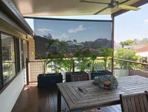 A wooden outdoor patio in Brisbane with a table, chairs, potted plants, a ceiling fan, and a mesh sunshade overlooks a suburban neighborhood.