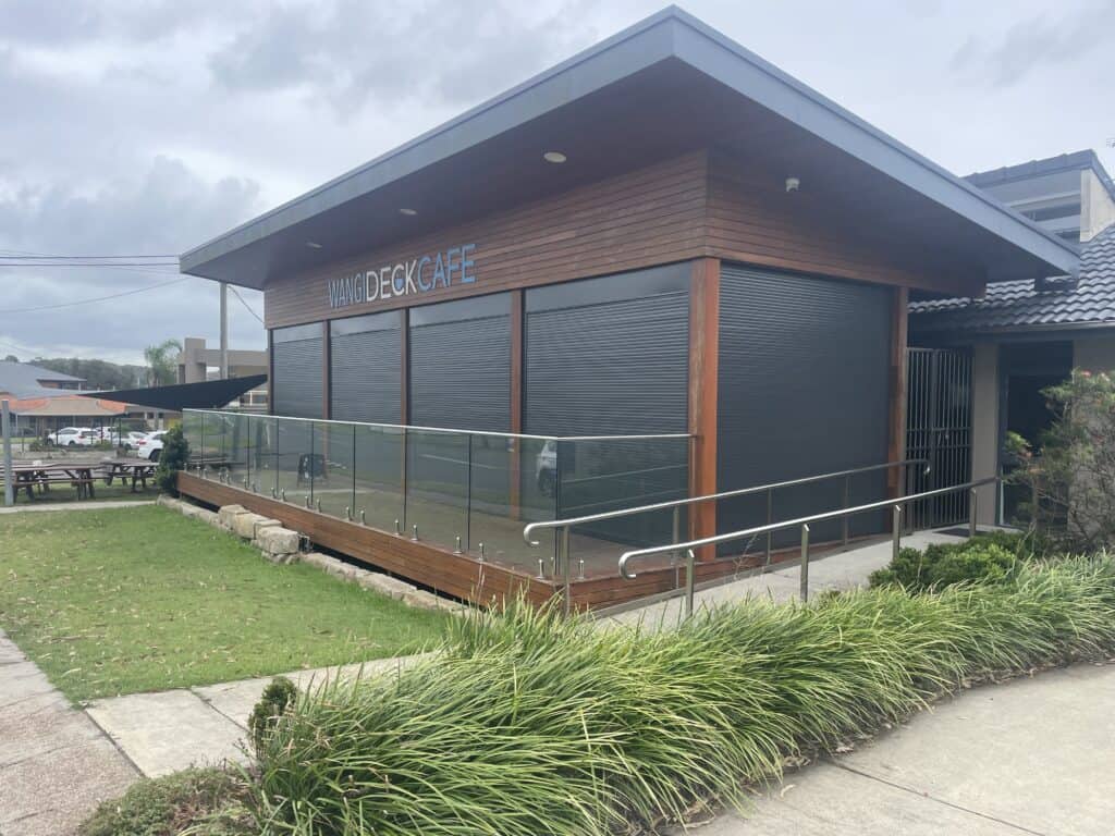 A modern café building with closed metal shutters, wooden exterior, glass railing, and a wheelchair-accessible ramp, surrounded by greenery under a cloudy sky.