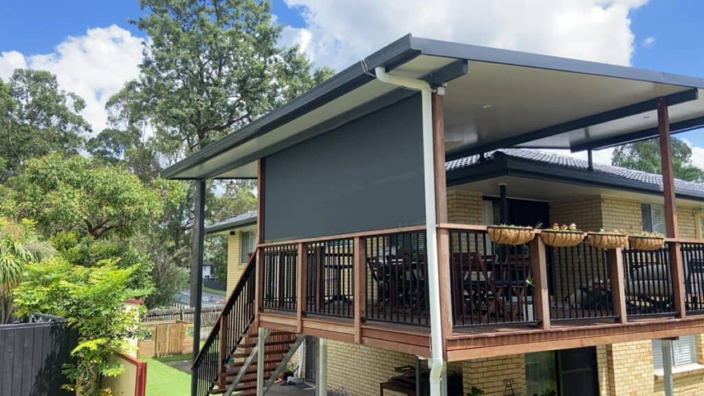 A raised patio with wooden railings and stairs features a large retractable outdoor blind and several hanging flower baskets, attached to a brick house in Brisbane under a cloudy sky.