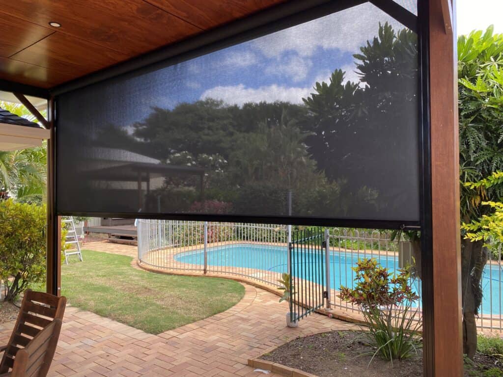 A backyard patio in Albany Creek with a black mesh shade, overlooking a fenced swimming pool, brick paving, and lush green plants under a partly cloudy sky.
