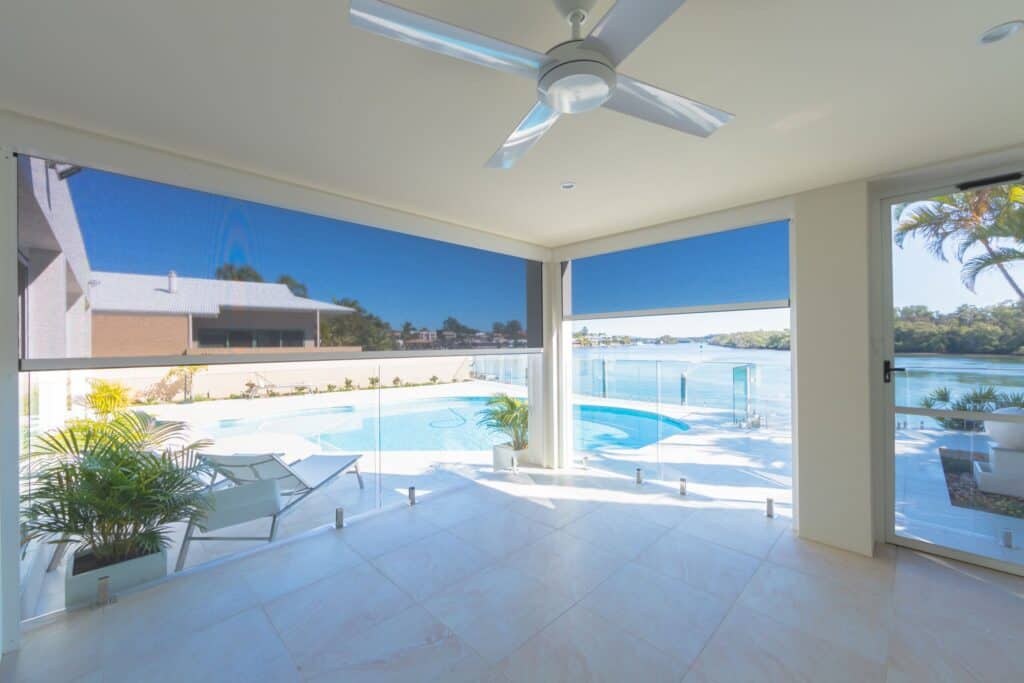 A spacious covered patio with ceiling fan overlooks a pool and picturesque Cleveland waterfront, featuring glass railings, loungers, and partial blue roller shades.