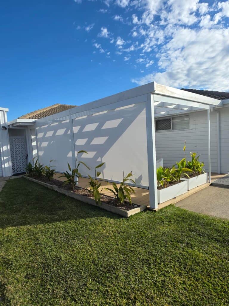 White pergola with planter boxes and green plants beside a white building, set on a lawn under a partly cloudy sky.