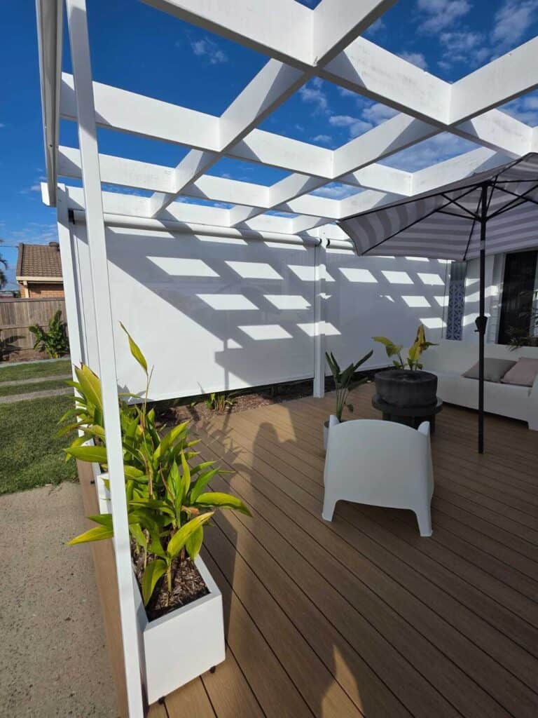 A modern outdoor patio with white pergola, wooden deck, potted plants, white furniture, and an umbrella under a partly cloudy sky.