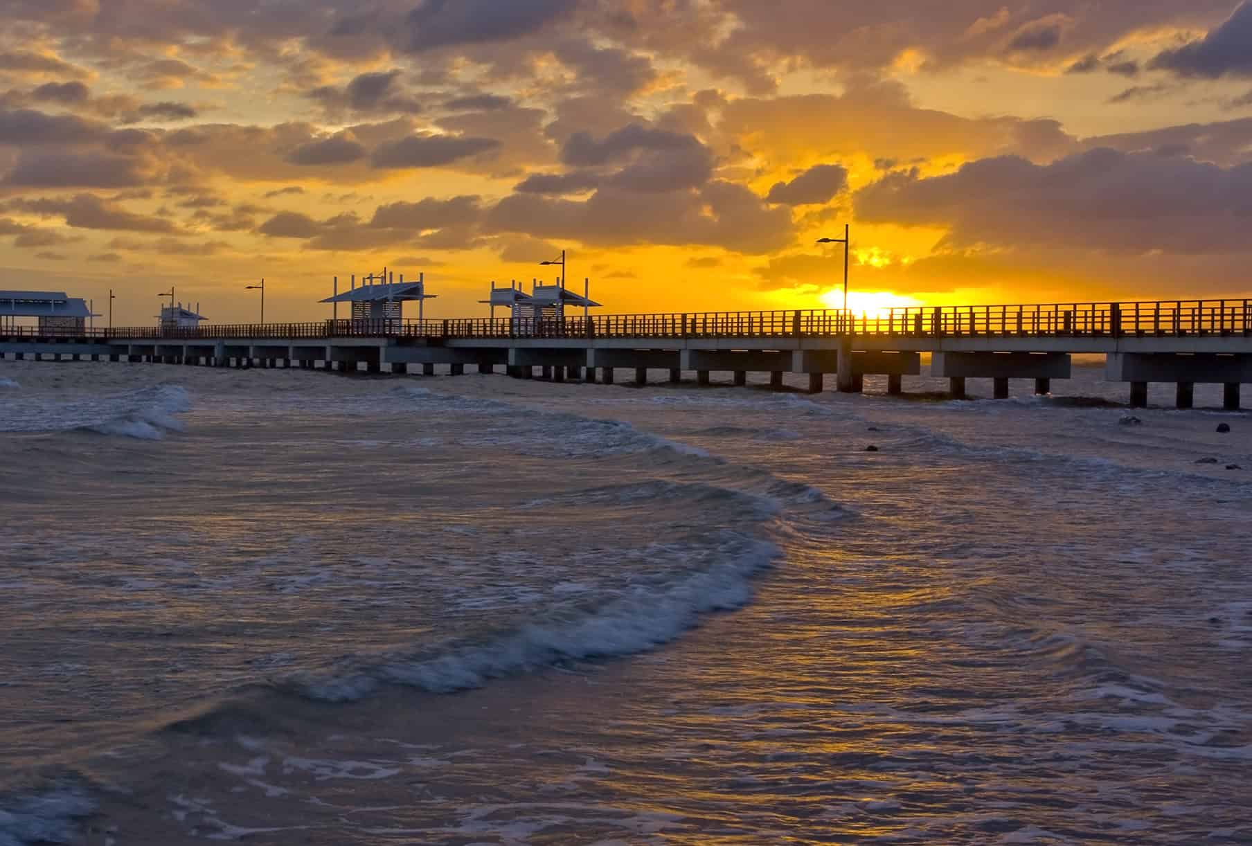A long, woody pier extends over the ocean at sunset, with waves in the foreground and clouds illuminated by the orange and yellow light in the sky.