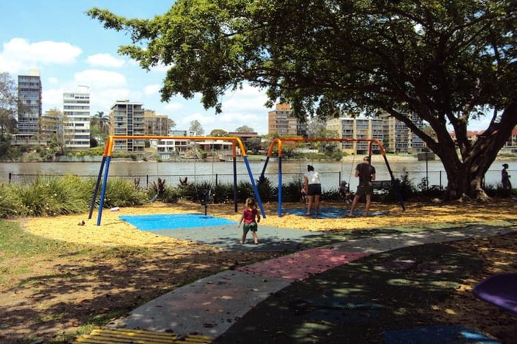 Children and adults enjoy a riverside playground with swings, shaded by a large tree, at Shailer Park, with apartment buildings visible across the water.