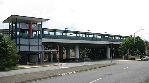 A modern railway station at Holland Park features an elevated platform, glass barriers, and an elevator tower, all visible from across a street with traffic lights.
