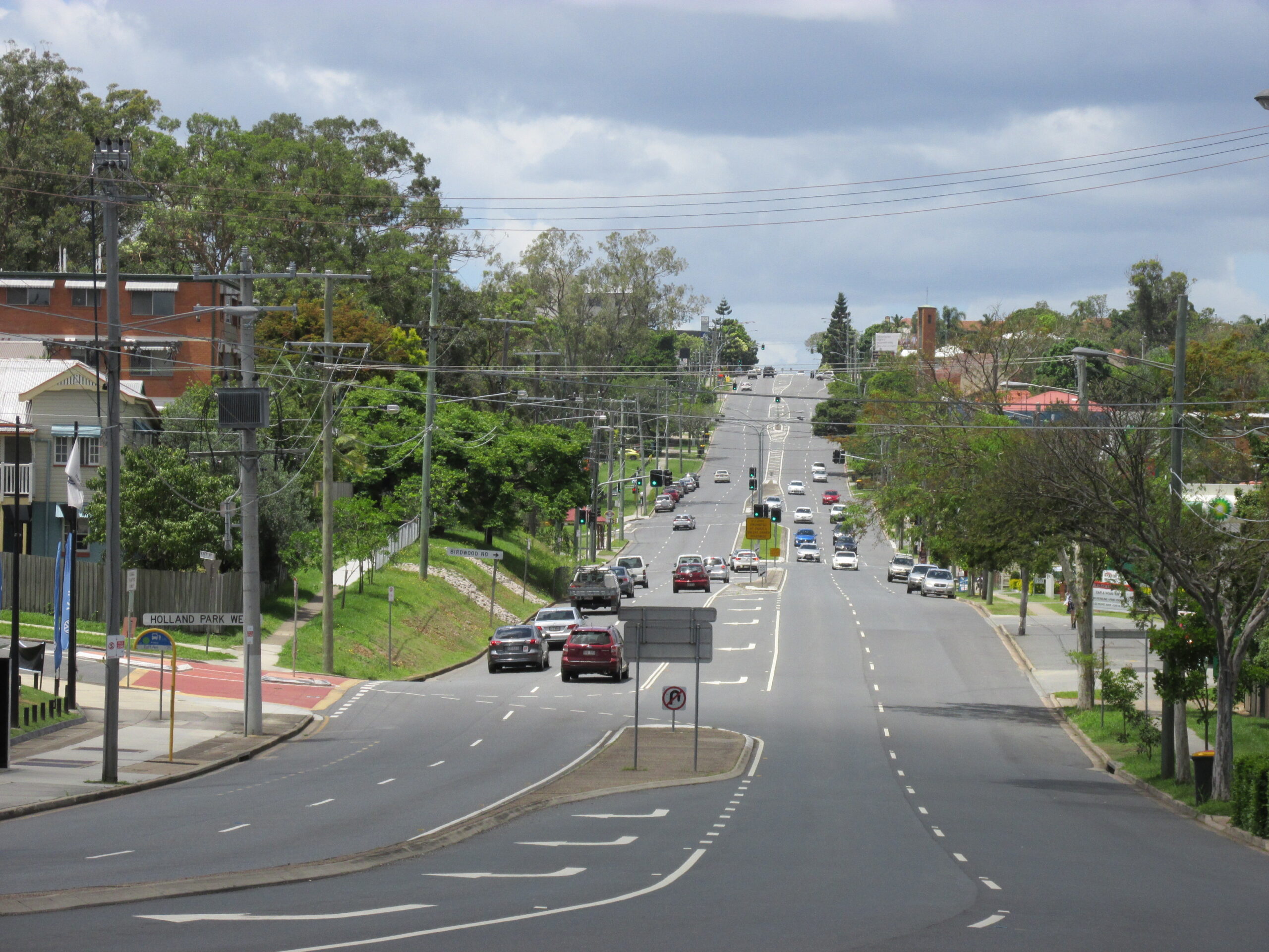 Wide, multi-lane suburban road in Holland Park with cars driving in both directions, lined with trees, power lines, and buildings under a cloudy sky.