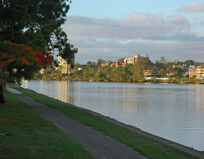 A paved riverside path in Shailer Park runs alongside calm water, with trees and buildings visible on the opposite bank under a partly cloudy sky.