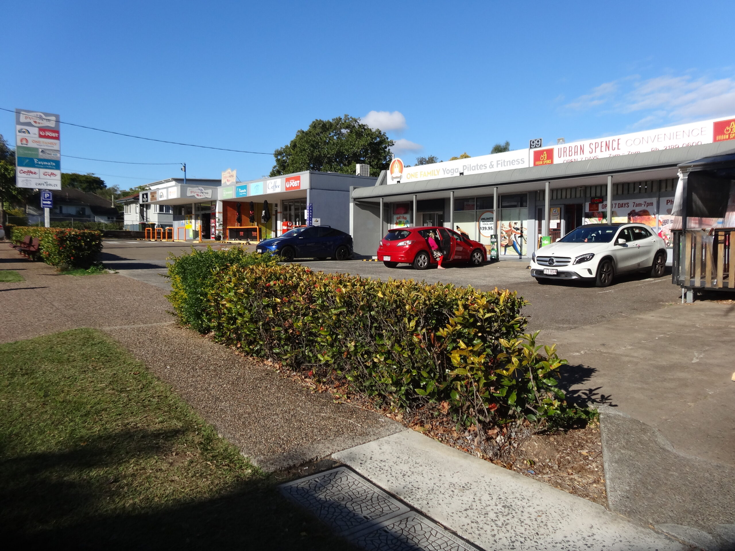 A small commercial strip in Holland Park with parked cars, storefronts, signs, and low hedges under a clear blue sky.