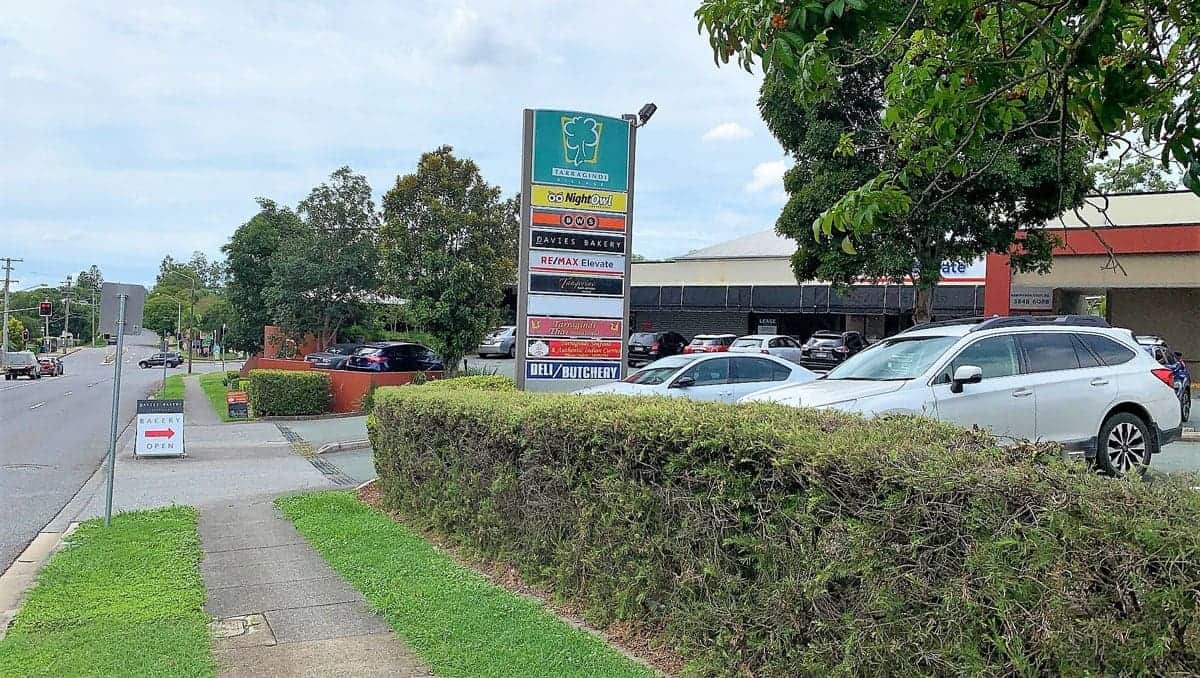 A shopping center sign in Shailer Park lists various businesses beside a parking lot with several cars; trees and a sidewalk line the street in the foreground.