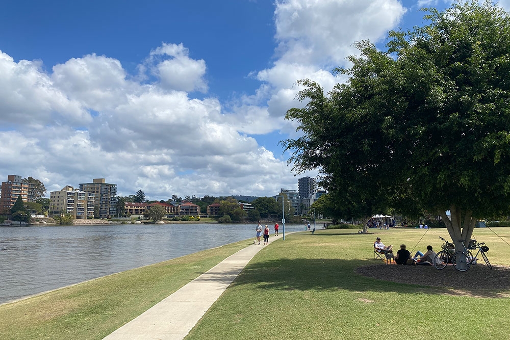 People walk and relax by a river on a sunny day in Shailer Park, with high-rise buildings in the background and a shady tree in the foreground.