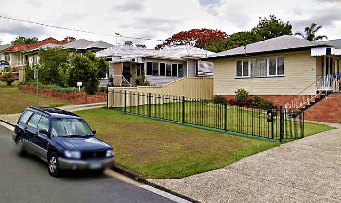 A blue station wagon is parked on the street in Shailer Park, in front of two single-story houses with neat lawns and a green metal fence.