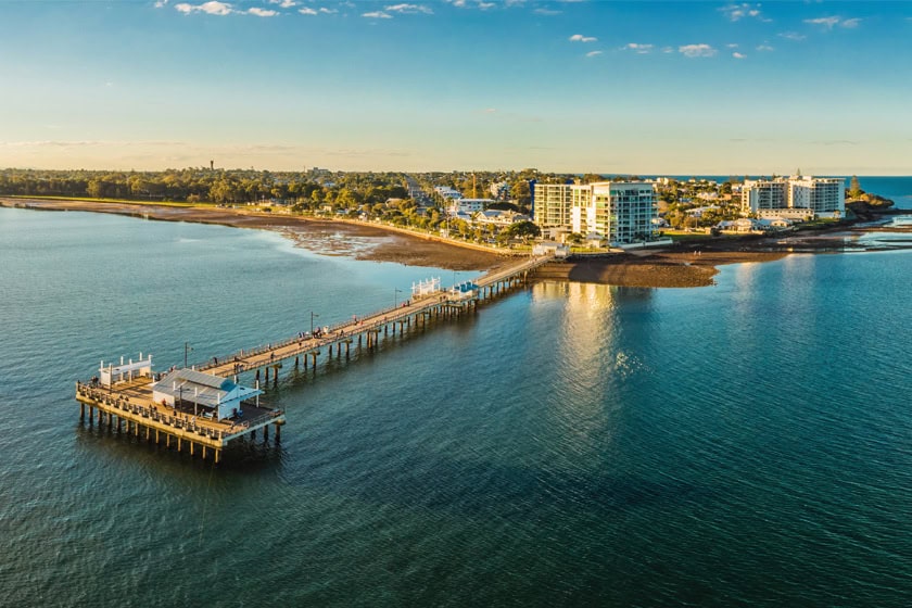 A long, woody pier extends over calm blue water toward a coastal town with buildings and trees under a clear sky.