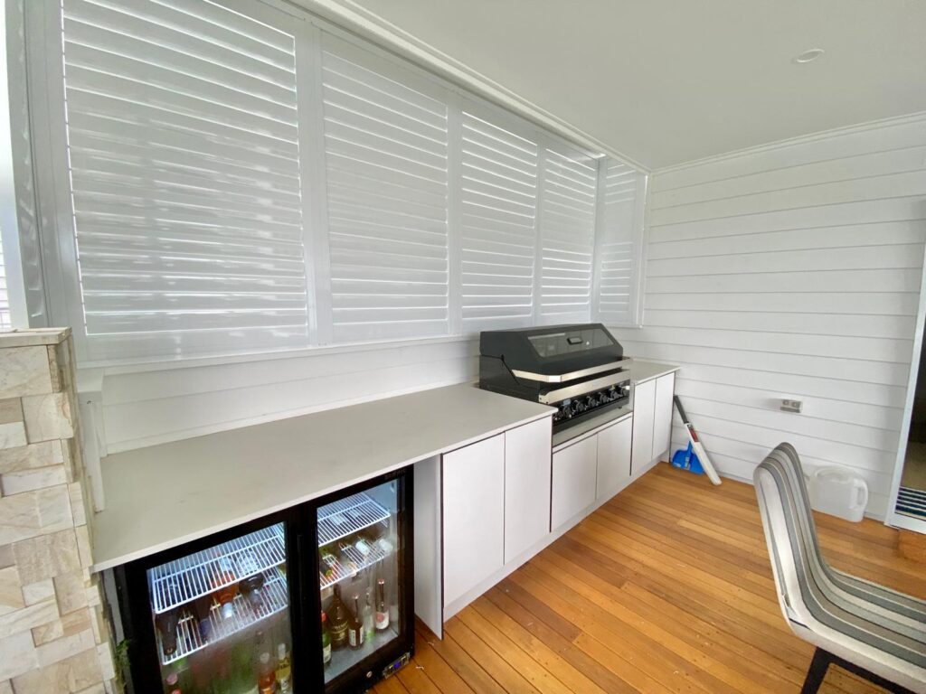 Outdoor kitchen area with a large grill, countertop, beverage fridge with drinks, white cabinets, and chairs on a wooden floor, enclosed by white shuttered windows.