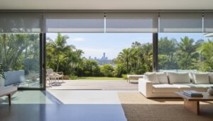 Modern living room with large glass doors open to a patio, outdoor chairs, lush greenery, and a distant city skyline under a clear sky.