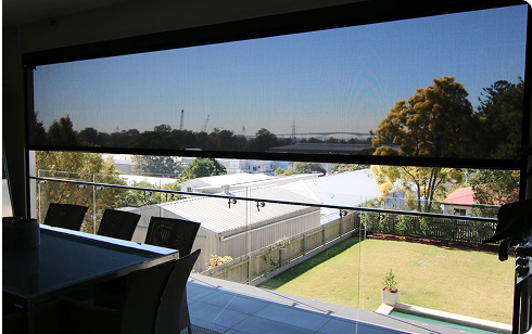 Outdoor view through a large glass window with a mesh roller blind, showing a patio, backyard, and trees under a clear sky.