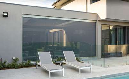 Two gray lounge chairs are placed beside a swimming pool in the backyard of a modern house with large windows and glass fencing.