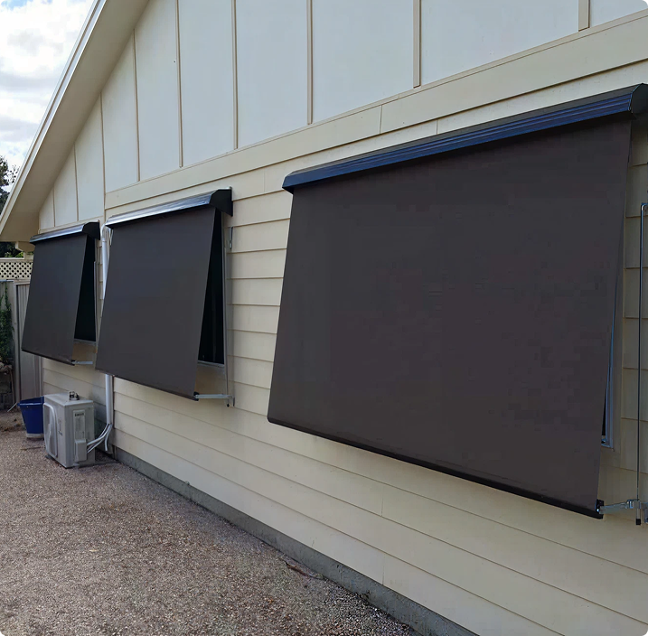 Three dark-colored awnings extend over windows on the side of a cream-colored house with horizontal siding.