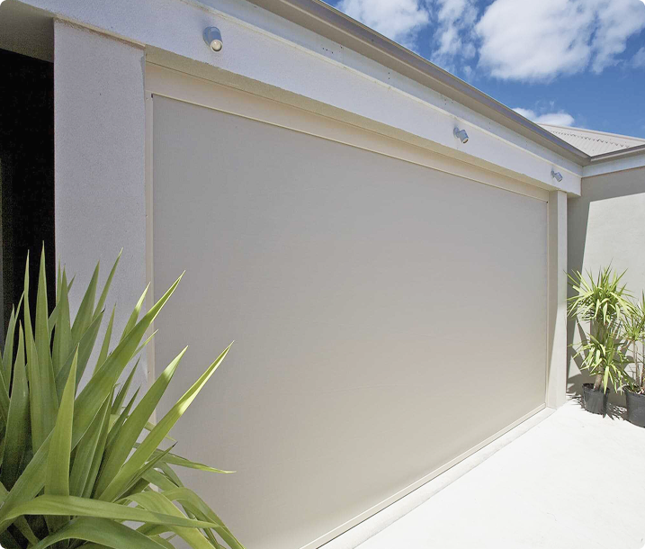 A closed beige roller garage door on a modern house, with green plants on either side and a partly cloudy sky above.