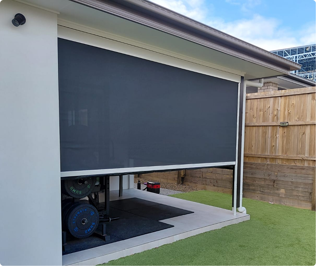 A covered outdoor gym area with a large black roller shade, weightlifting equipment inside, and a wooden fence in the background.