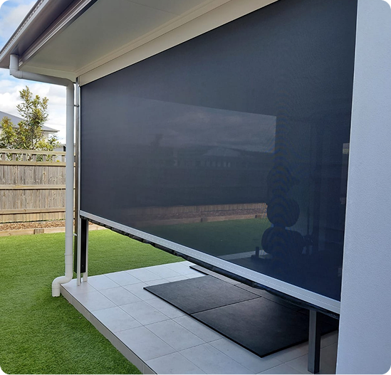 A covered patio with a dark outdoor roller blind, artificial grass lawn, wooden fence, and a small mat near the door.