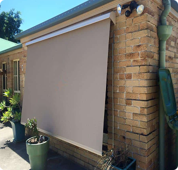 A tan outdoor roller blind partially lowered over a window on a brick house, with potted plants nearby and a green downspout attached to the wall.