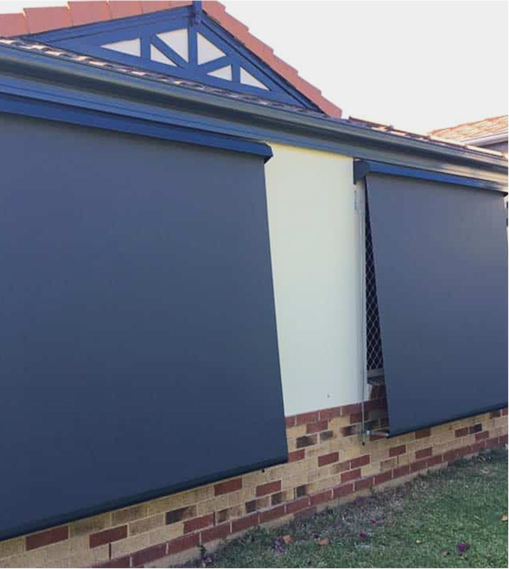 Two large outdoor roller blinds cover windows on a brick and cream-colored house with a gabled roof.